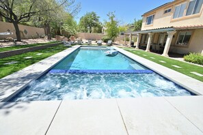 Una piscina rectangular con un jacuzzi, rodeada por una terraza con sillas y una zona de estar cubierta.