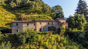 Un bâtiment en pierre, doté d’un toit en tuiles, entouré d’une végétation luxuriante et d’arbres.