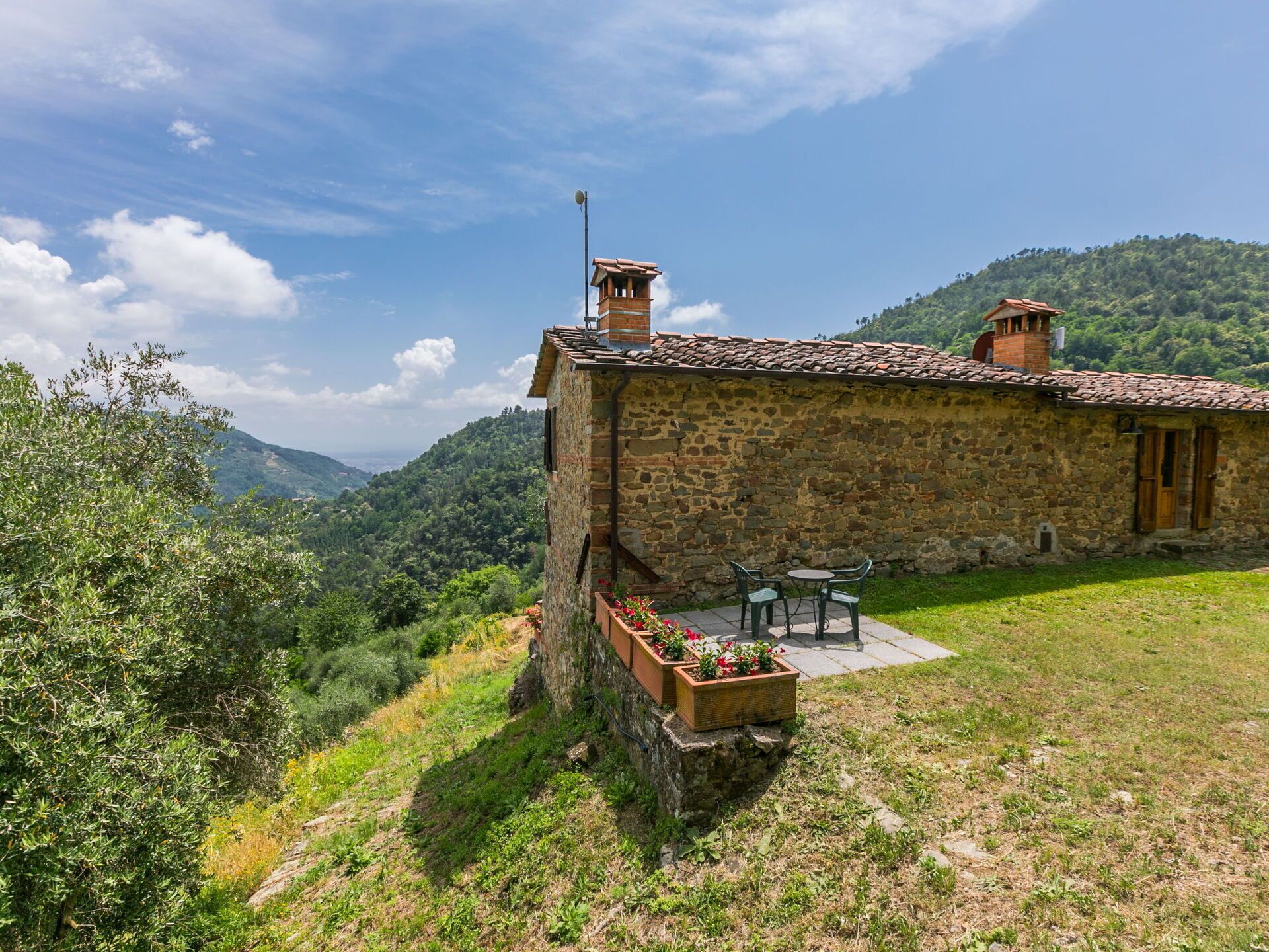 Una casa de piedra con techo de tejas, un pequeño patio con mesa y sillas, y vista a un valle exuberante.