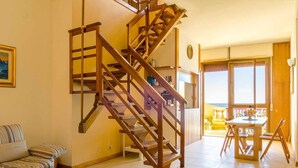 A spacious living area with a wooden staircase, a sofa, a dining table with chairs, and a view of the sea through large windows. - La Plage - Home and Sea (Nizza Di Sicilia)