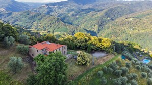 Un edificio in pietra con tetto rosso, circondato da vegetazione lussureggiante e colline sullo sfondo.