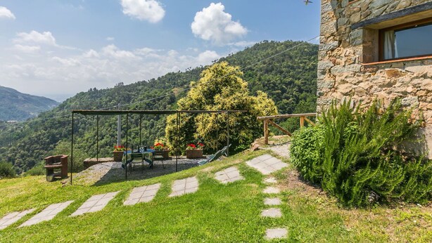Un edificio in pietra con un'ampia finestra, un giardino con tavolo e sedie, e vista su una montagna boscosa.
