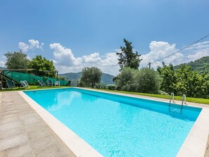 Ein rechteckiger Pool mit klarem, blauem Wasser, umgeben von einer Steindeck. Liegestühle und eine Pergola mit grüner Markise sind vorhanden. Im Hintergrund eine üppige, grüne Landschaft mit Bäumen und Hügeln unter blauem Himmel.