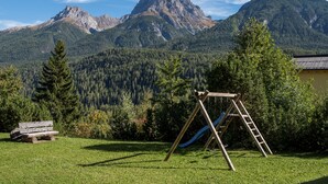 Eine Wiese mit einer Holz-Schaukel und einer Kletterleiter, umgeben von Bäumen und einem Berg im Hintergrund.