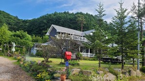A traditional wooden house with a white porch, surrounded by lush greenery and a clear sky. - Under Hill Pension (Pyeongchang)