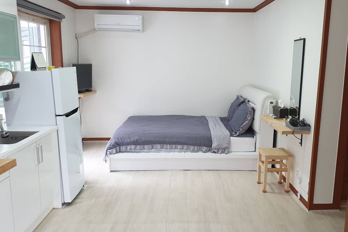 A compact room with a bed, a small stool, a desk with a mirror, a shelf, and a window with a calendar.