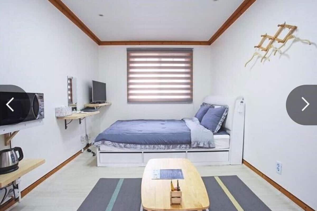 A compact bedroom with a bed, a desk, a television, and a window with blinds.