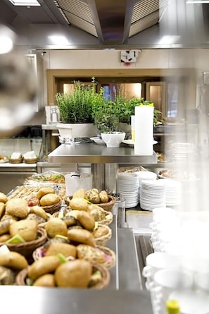 A commercial kitchen with stainless steel surfaces, potted plants, and food items like mushrooms and bread.