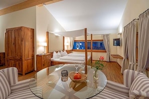 A hotel room with a bed, a glass table with a bowl and wine glasses, a wooden wardrobe, and a TV on the wall.