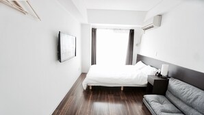 A modern hotel room with a large bed, a grey sofa, a bedside table, and a mounted TV. - Hostel 758 Nagoya 4P (Nagoya)