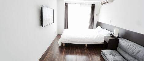 Una habitación de hotel moderna con una cama grande, un sofá gris, una mesita de noche y un televisor montado.