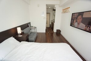 A hotel room with a bed, a nightstand, a lamp, a gray sofa, and a television screen displaying a program. - Hostel 758 Nagoya 4P (Nagoya)