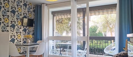 A room with a dining table, chairs, a hanging light fixture, a sofa, and a view of the outdoors through large windows.