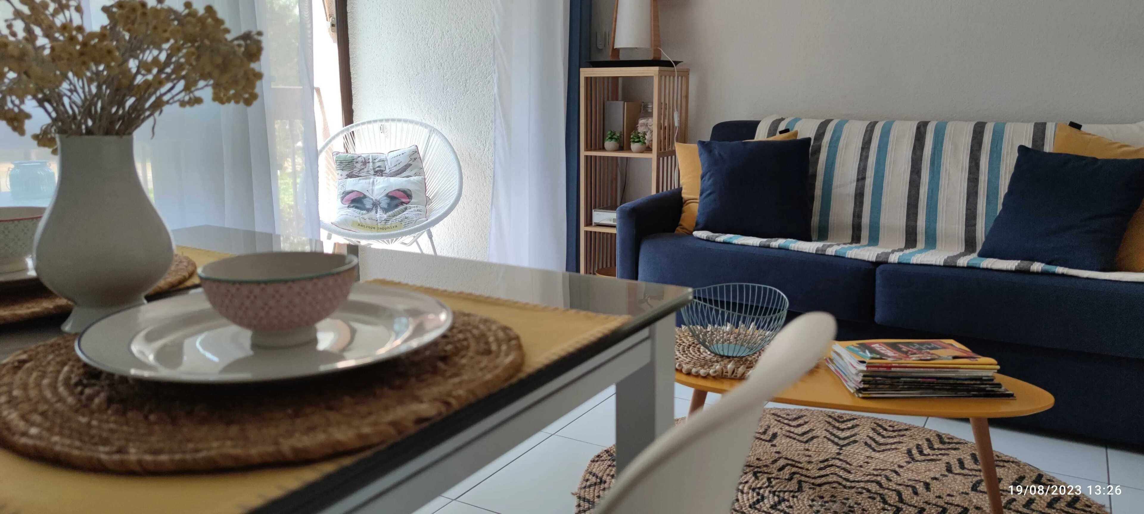 Ett vardagsrum med en soffa, ett soffbord, en fåtölj, en bokhylla och en vas med torkade blommor.