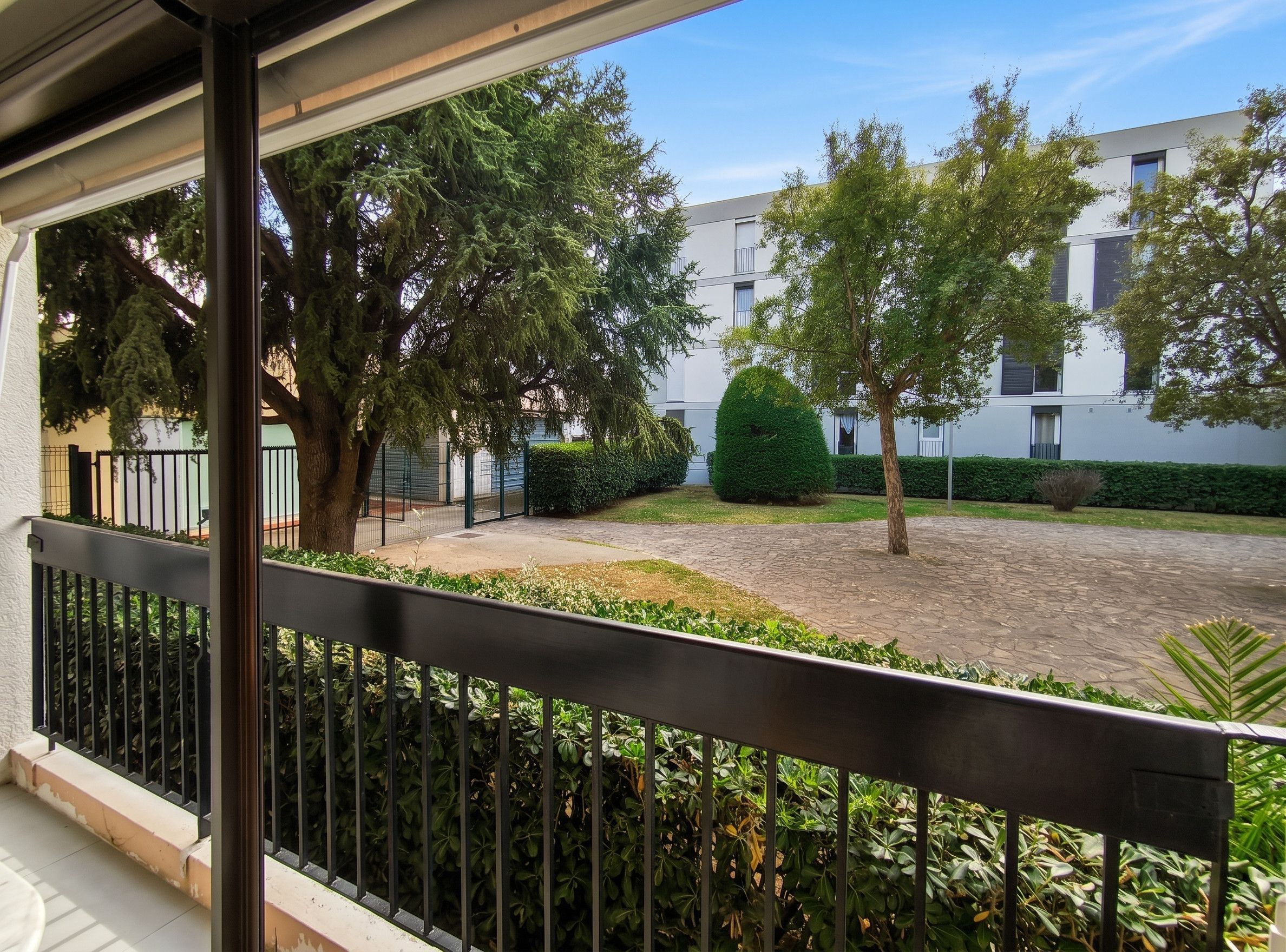 Ein Balkon mit Blick auf einen Innenhof, ein Gebäude mit Flachdach und ein gepflegter Heckenstreifen.