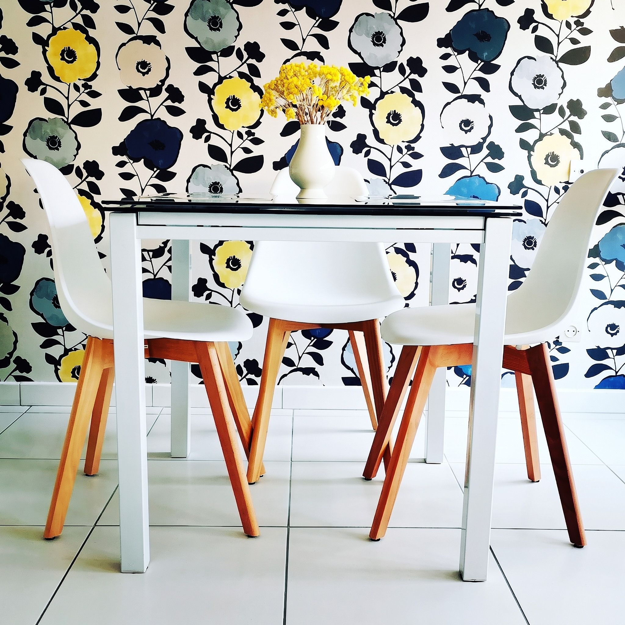 Une table à manger avec quatre chaises, un vase rempli de fleurs jaunes et un papier peint à motifs floraux.