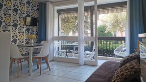 Une pièce comprenant une table à manger, des chaises, un canapé, un téléviseur et un balcon donnant sur des arbres et une clôture.