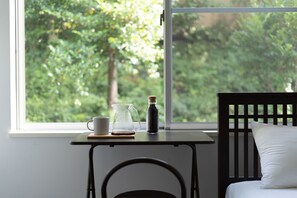 A room with a window overlooking a forest, a small table with a cup, a pitcher, and a bottle. - Satellite Hotel Yebisu (Tokyo)