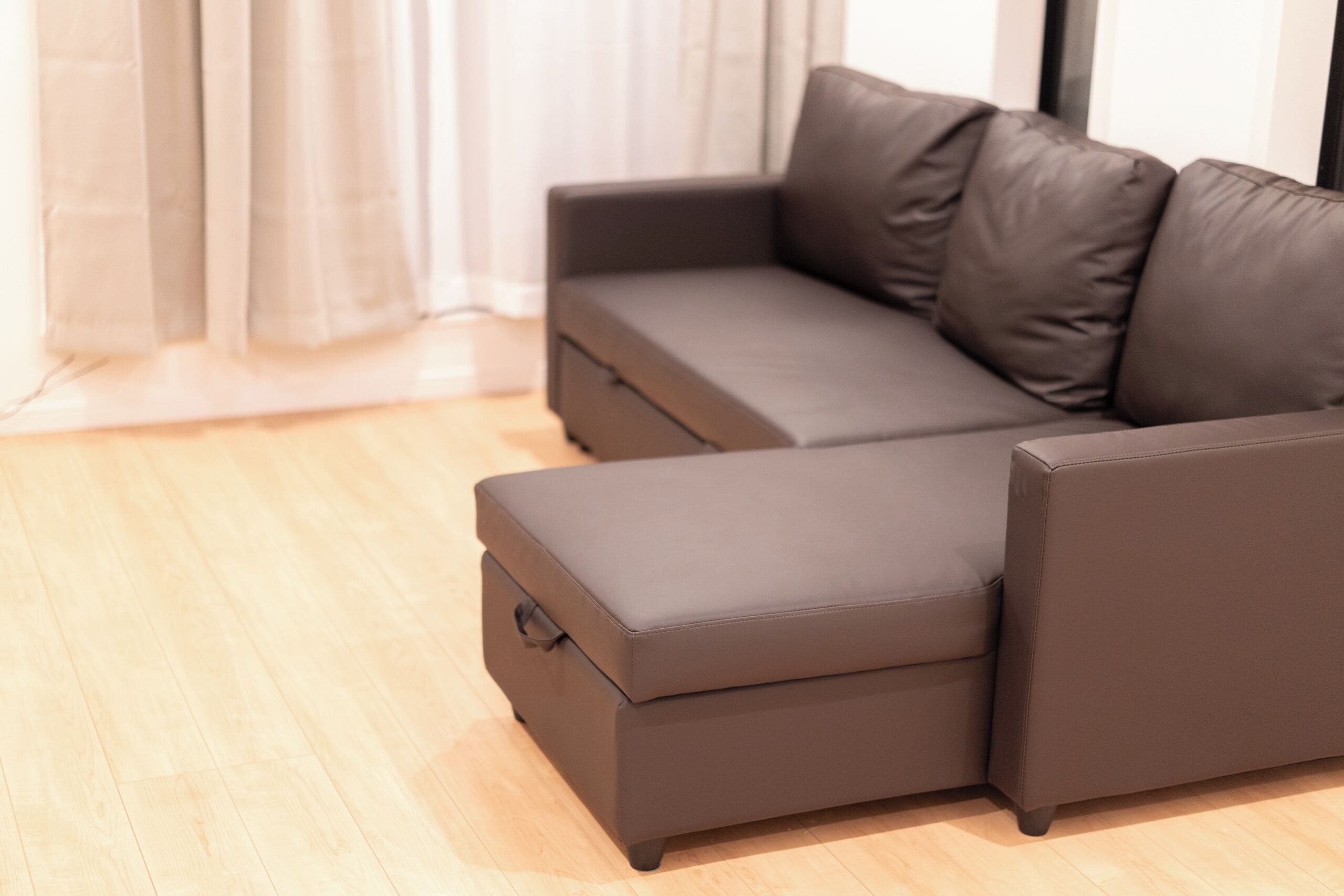 A modern living room with a brown leather sofa and ottoman, light wooden flooring, and a window with sheer curtains.