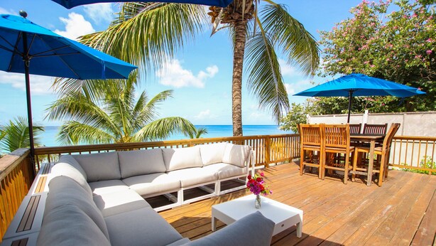 Een houten terras met een witte bank, houten stoelen en twee blauwe parasols.