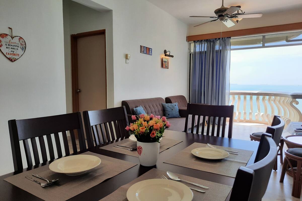 Un coin repas avec une table dressée pour quatre personnes, un ventilateur de plafond et une vue sur la mer depuis un balcon.
