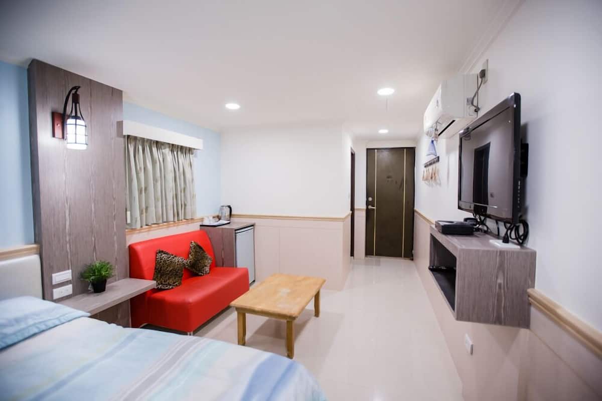 A compact hotel room with a bed, a red sofa, a small table, a TV, and a kitchenette.