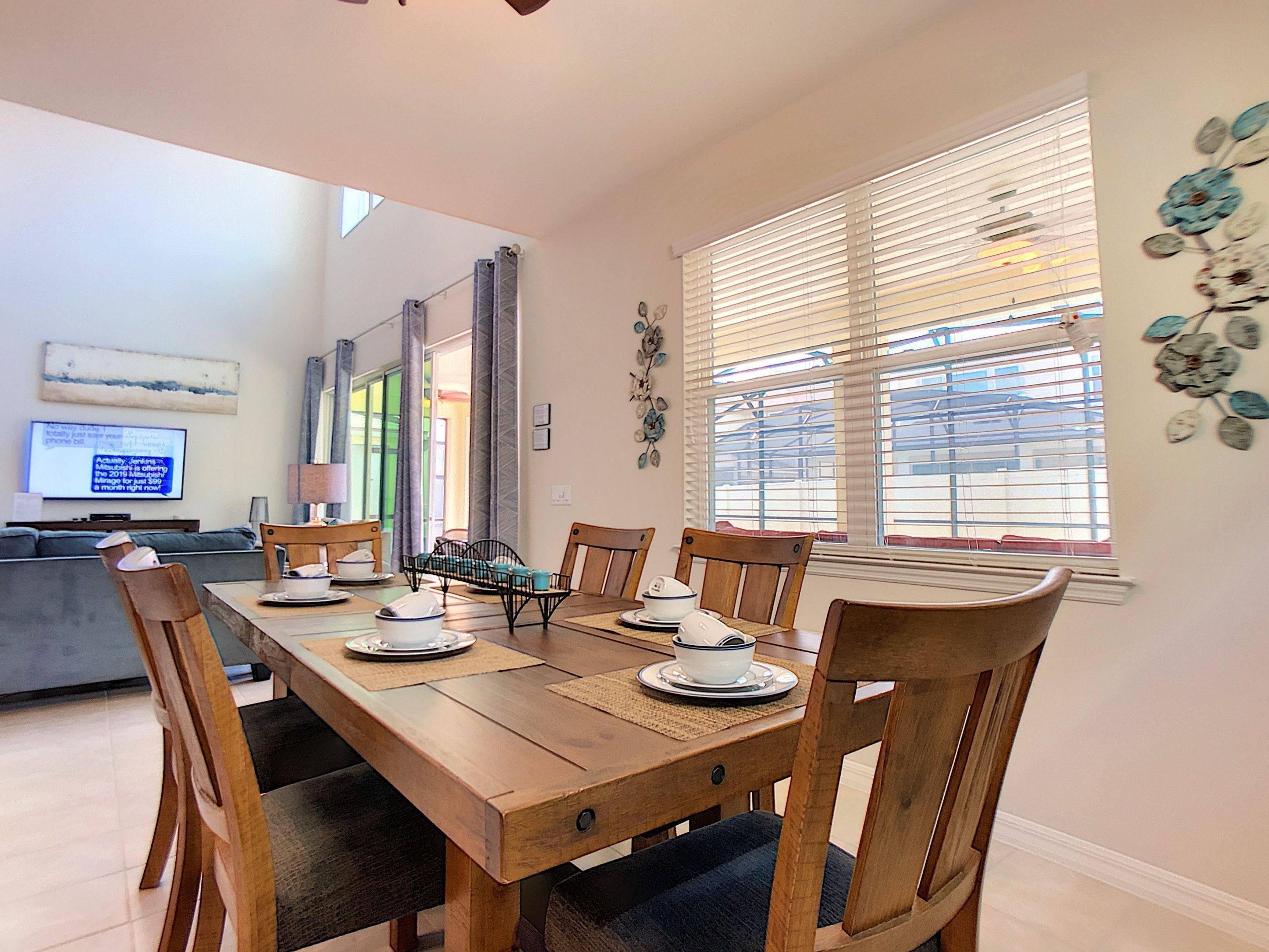 A dining area with a wooden table set for six, surrounded by chairs. A television is mounted on the wall in the background.