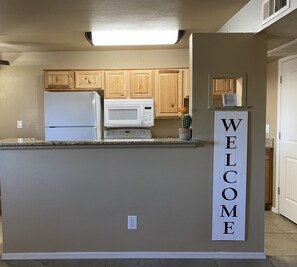 A kitchen with wooden cabinets, a white refrigerator, a microwave, and a welcome sign.