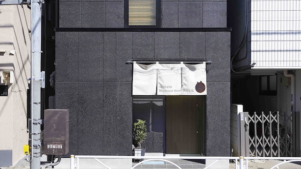 La façade d’un immeuble porte une banderole sur laquelle on peut lire « IKIDANE, hôtel résidentiel », et une petite plante en pot est placée près de l’entrée.