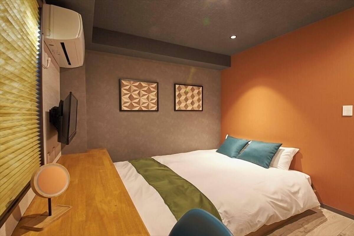 A hotel room with a bed, a desk, a chair, two wall-mounted televisions, and a door.