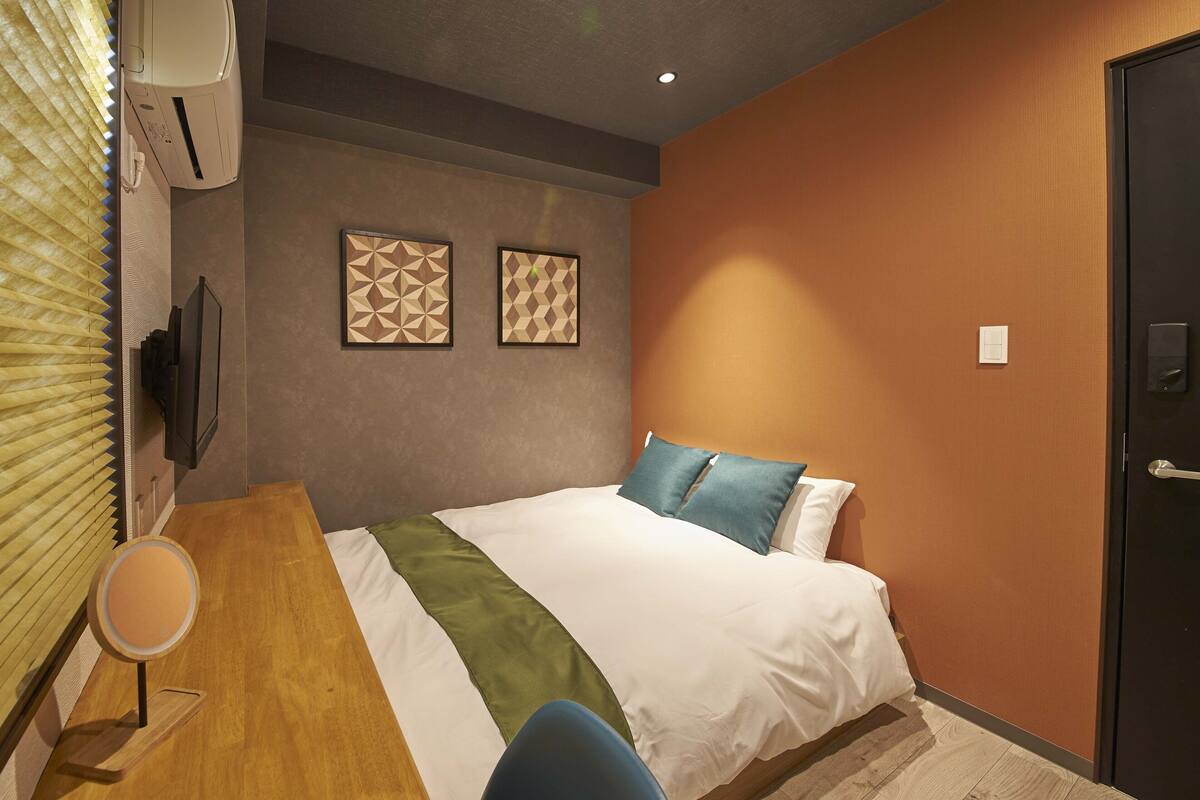 A hotel room with a bed, a desk, and a chair. There are two wall-mounted air conditioning units and two framed geometric wall art pieces.