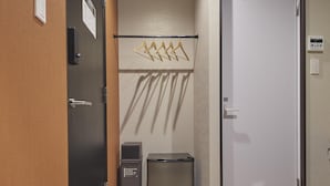 Un couloir avec une porte en bois, un porte-manteau avec des cintres, une poubelle et un thermostat fixé au mur.