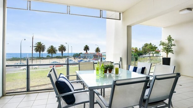 Un comedor moderno con una mesa y sillas de vidrio, con vista a una playa con palmeras y cielo despejado.