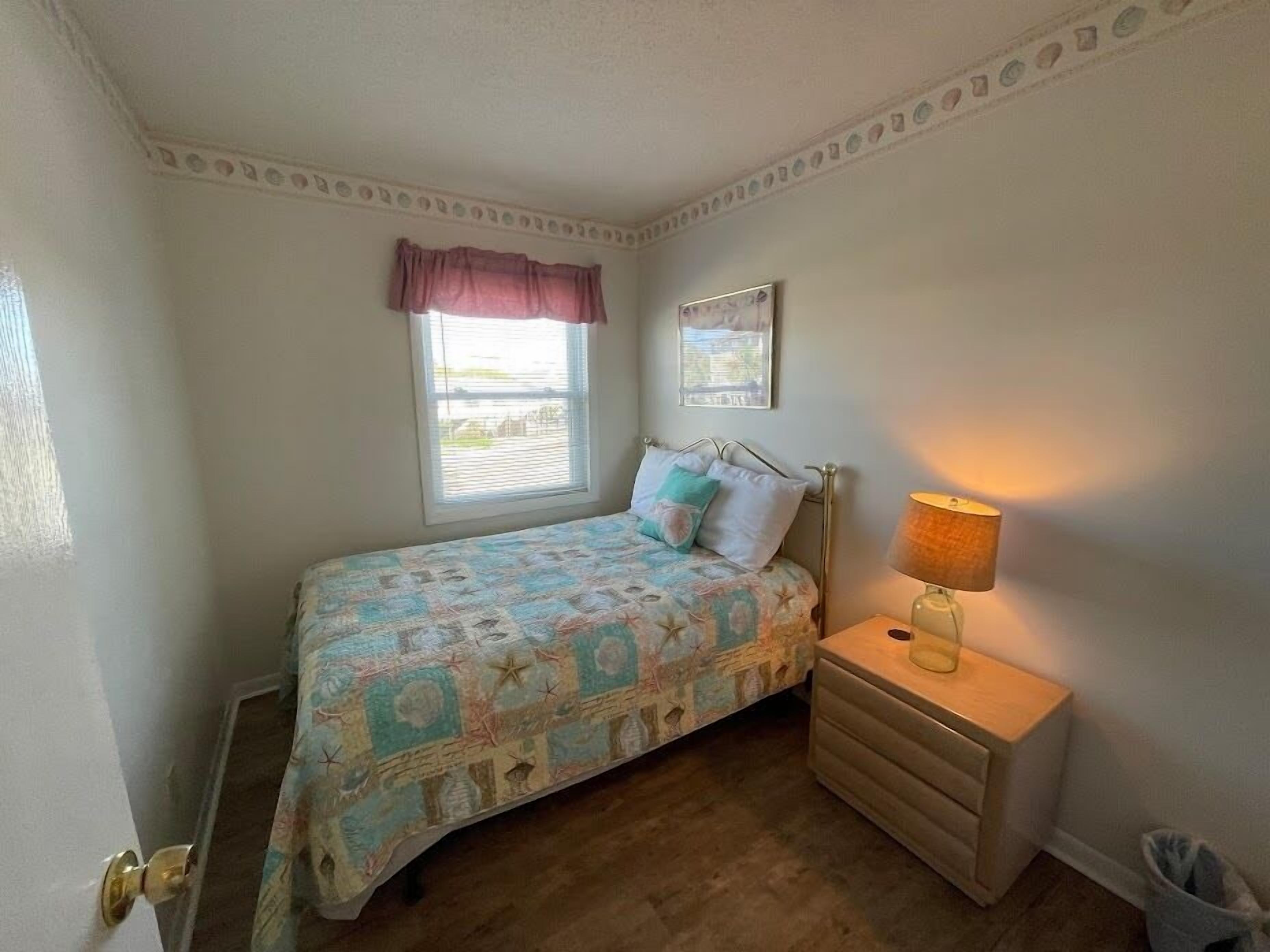 A bedroom with a bed, a bedside lamp, a window with blinds, and a small picture on the wall.
