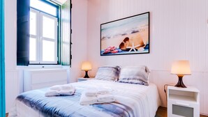 A bedroom with a bed, two bedside lamps, a framed picture of a beach scene, and a window with blinds. - TREM HOUSE - LOCATED IN OLD CITY - FARO (Faro)