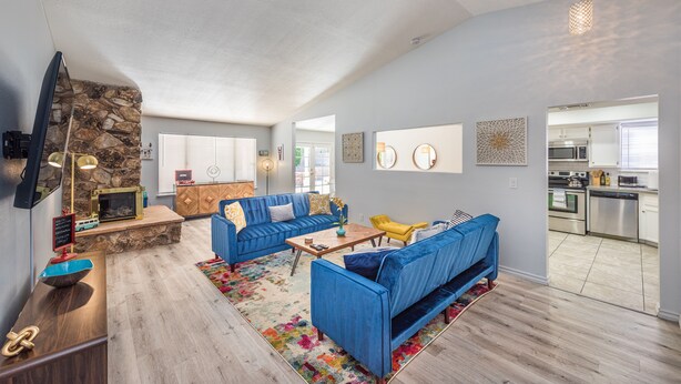 A modern living room with a stone fireplace, blue sofas, a coffee table, and a kitchen in the background. - Charming & Stylish home can accommodate 8 guests! (Henderson)