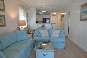 Uma sala de estar com um sofá azul, uma mesa de centro e uma cozinha ao fundo.
