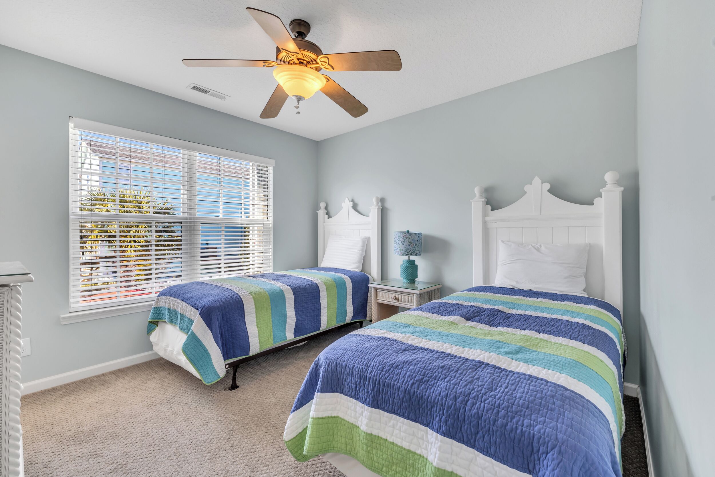 A bedroom with two beds, a ceiling fan, a window with blinds, and a nightstand with a lamp.