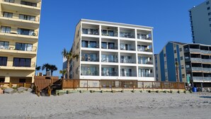 Een hotel met meerdere verdiepingen en balkons, gelegen aan een zandstrand met palmbomen.