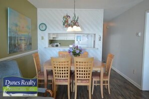 Un coin repas avec une table et des chaises, une horloge au mur et une cuisine en arrière-plan.