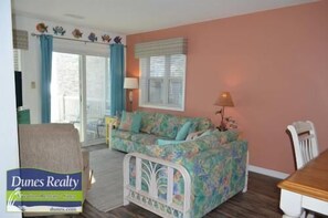 A living room with a floral sofa, a white baby crib, a dining table, and a window with blinds.