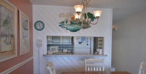 A dining area with a wooden table and white chairs, a kitchen area with white cabinets and appliances, and a wall-mounted clock.