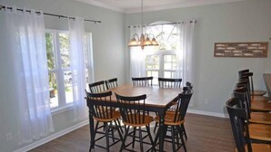 A dining room with a wooden table and chairs, large windows with white curtains, and a chandelier. - Bikini Bottom - Walk to Beach in Surfside Beach (Surfside Beach)