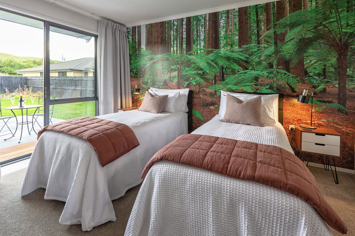 A bedroom with two beds, a large window, and a forest mural on the wall.