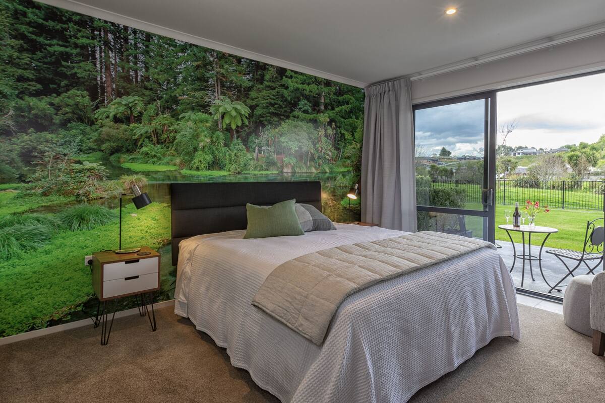 A bedroom with a large bed, a nightstand, a lamp, and a view of a garden through a glass door.