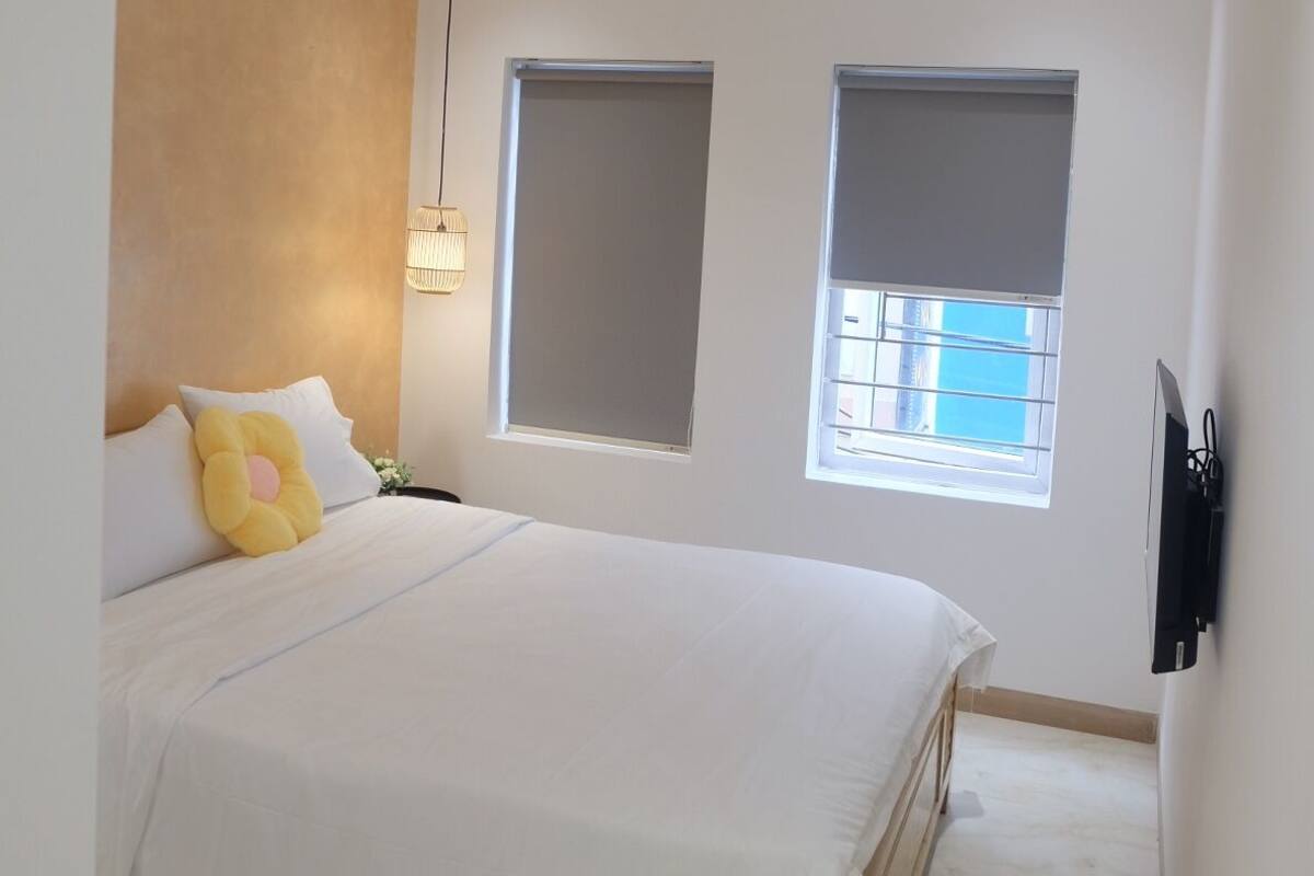 A modern bedroom with a large bed, two windows with blinds, a ceiling light, and a wall-mounted TV.