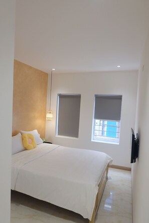 Un dormitorio moderno con una cama grande, dos ventanas con persianas, una lámpara de techo y un televisor montado en la pared.