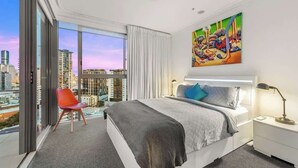 Une chambre d’hôtel moderne avec un grand lit, un fauteuil rouge, une vue sur la ville et un tableau coloré accroché au mur.
