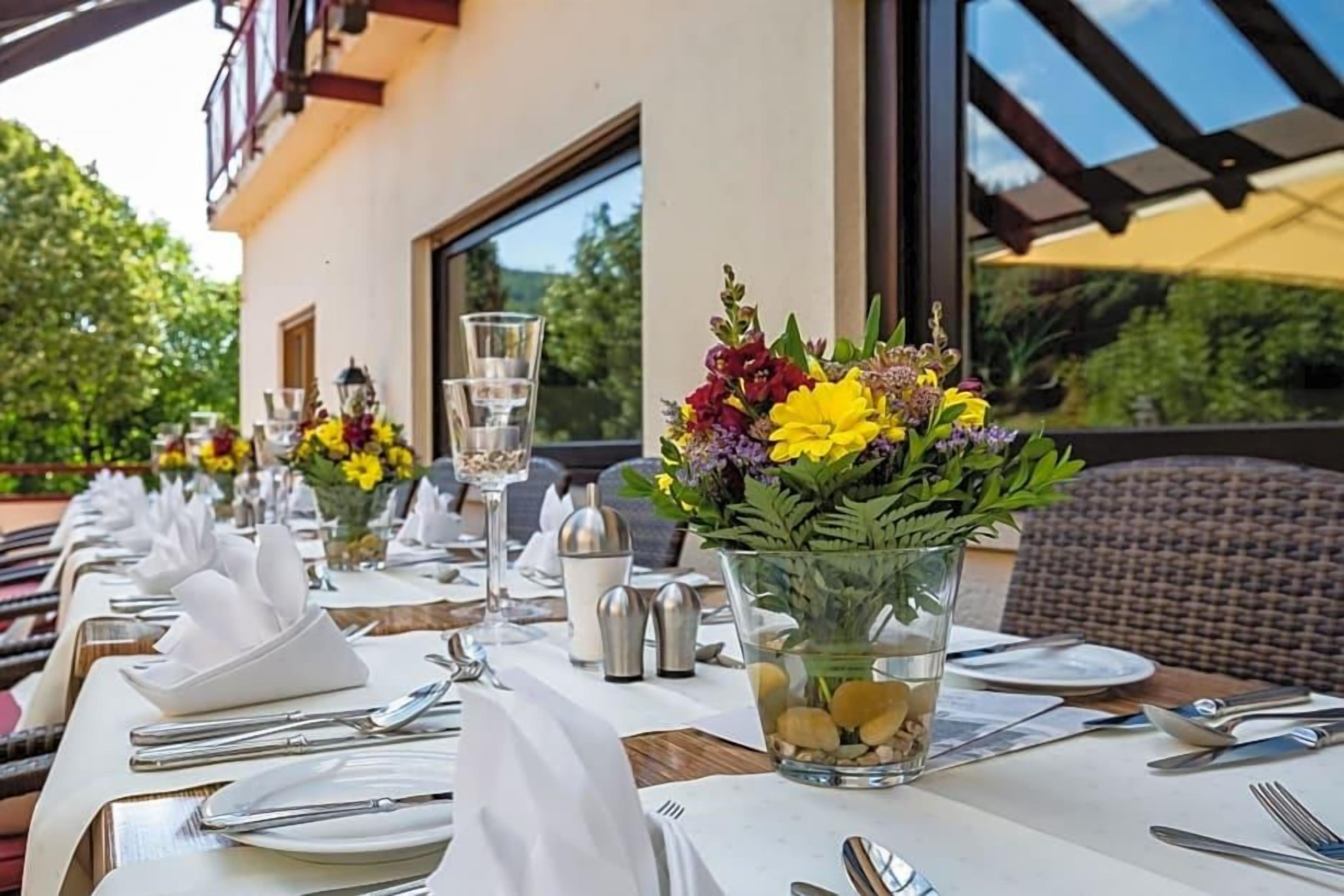 Ein Esstisch ist mit weißen Tischdecken, Besteck und Gläsern gedeckt, mit einer Blumenvase und einer Wasserkaraffe darauf.