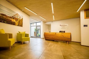 Zona de recepción con un mostrador de madera, dos sillas amarillas y un cuadro grande con un paisaje.